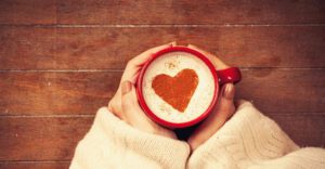 A mug of coffee with a red heart in the foam. The is held by a woman