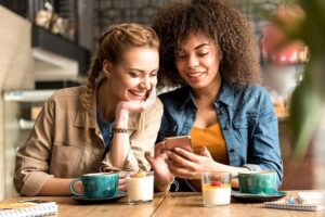 Two girls in a coffee shop laughing about something they see on their phone.