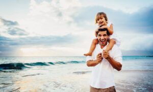 A happy dad with a his daughter on his shoulders at the beach.
